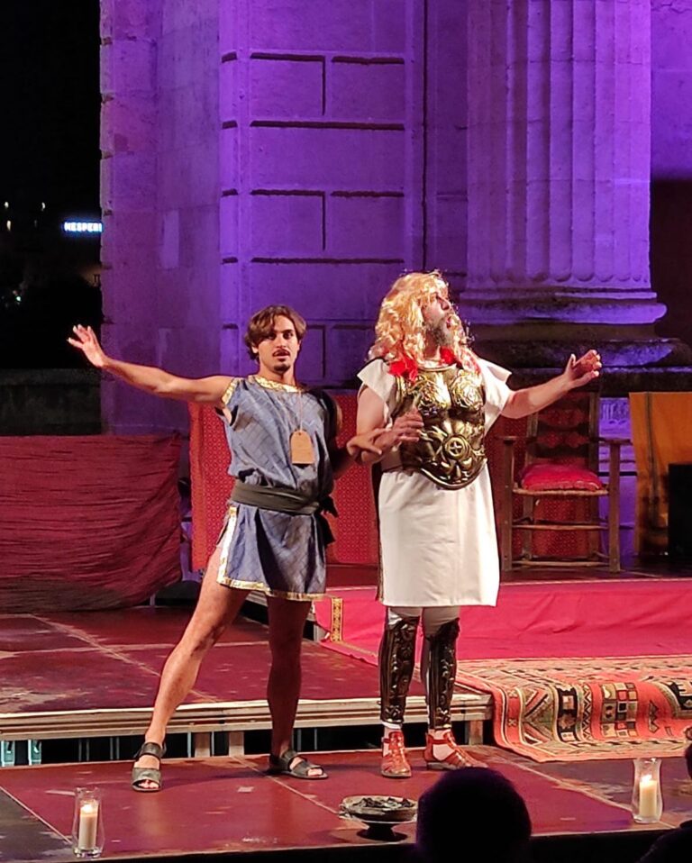 Actores representando la obra Miles Gloriosus: dos hombres con vestimenta romana, en pose de cantar o recitar y bailar, sobre fondo de columnas romanas e iluminación colorida.